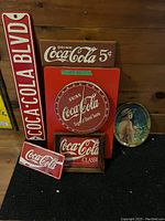 Overview of Coca-Cola collectible signs and thermometer arranged against a wood background, showing the full set.