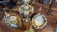 Photo showing three ornate gold painted Italian china pieces with floral decoration: a covered dish with handles, an urn or vase, and a scalloped bowl.