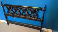 Full view of black wooden headboard with carved scrollwork against blue wall and carpeted floor.
