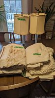 Photo shows two beige fabric table lamps with decorated shades and a stack of folded tablecloths and linens on a dining table under natural light.