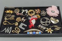 Top-down photo showing a box containing a collection of 23 assorted costume brooches in various designs, materials, and colors.