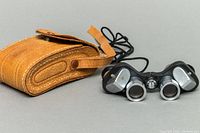 Baker Standard 6x15 opera glasses with attached neck cord beside tan leather carrying case resting on a gray background.