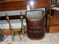 Full view of metal Ken-More ash bucket and pair of brass andirons on carpeted floor in front of wooden bench.