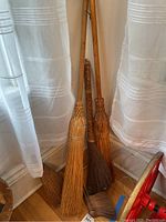 Five brooms leaning against a wall near curtains, including three long-handled decorative brooms and two smaller hand whisk brooms with natural straw and twine bindings.