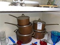 Four copper pots stacked on a shelf, two with loop handles and two with long handles, lids visible on top couples.