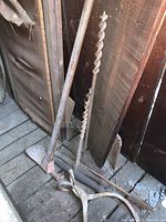 Photo showing long-handled post hole digger, hand auger drill, and possibly parts of other tools leaning against wooden wall.