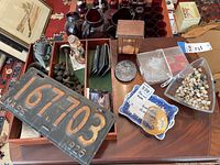 Vintage license plate, glass container with stones, plaques, bottle opener, wood box, rosary and zipper styled wallet arranged on a wooden table.