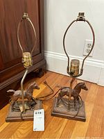 Two horse base table lamps without lamp shades, showing metal harps and wooden rectangular bases with horse figures.