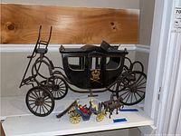 Wide view showing the large black royal carriage style coach wagon alongside the smaller blue coach with horses and figurines, placed on a white shelf.