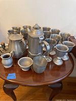 Full set of pewter goblets, teapot, decanter, creamer, chalices, and candlestick holder arranged on wooden table.