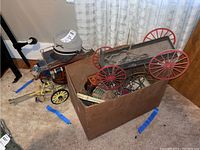 Full view of box containing parts including red wheeled covered wagon frame, miniature accessories in box, and metal wagon with barrel seen in background