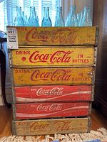 Front stacked view of six wooden Coca Cola crates with visible yellow and red Coca Cola branding and paint wear.