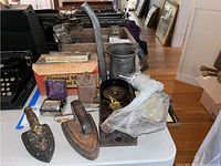 Table display of two vintage irons, Rolls Razor in box, a metal pitcher, Tork clock, and small metal bank. Various items show surface rust and wear.