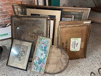 Several framed artworks and empty frames leaning against a wall on the floor, showing vintage prints and water-damaged frames.