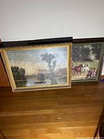 Two framed paintings leaning against a white wall on wooden floor, showing a river landscape and a horse carriage with figures.