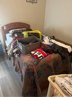 Single wood bed with headboard and footboard, mattress with plaid pillowcases, patchwork quilt, various bags and other items on bed surface.
