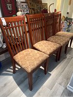 Four wood dining chairs with vertical slat backs and patterned beige upholstered seats placed in a row on a wood floor.