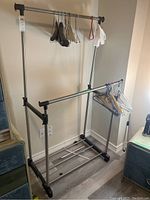 Full front view of metal rolling clothes rack with various hangers and plastic clips attached, standing on laminate flooring near wall.