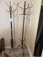 Photo of two coat stands, one metal with brass finish and one wooden, both standing side by side on floor.