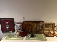 Overview photo of all items on a cabinet top against a white wall, showing the copper diorama, three framed prints, hurricane lamp, glass and ceramic decor items.