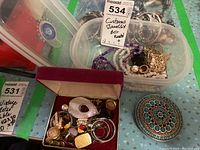 Overview of a plastic container with assorted costume jewelry including bead necklaces (white and purple), a red velvet box with earrings and pins, and a round belt buckle with turquoise-colored beads.