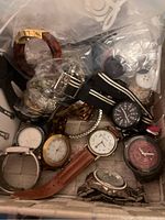 Wide view of the box containing multiple watches and bracelets including metal, leather, and plastic bands along with assorted jewelry pieces in plastic bags.