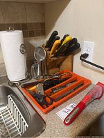 Full view of kitchen counter with orange tray containing knives and scissors, wooden knife block behind with black and yellow handled knives, paper towel holder, and metal strainer visible