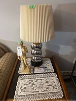 Shows full view of statue and lamp on a dark wood table with lace mats, highlighting size and condition