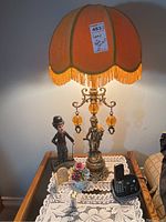 Full view of the lamp with orange fabric shade, metal figurine base, and Charlie Chaplin figurine next to it on table.