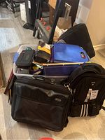 Photo showing a collection of office supplies including black briefcase, black and blue knapsacks, folders, notebooks, and staplers piled in a plastic container and on the floor.