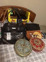 Overview of purses including black purse with braided handles, tan purse with dog and moon print, and two decorative round metal boxes with intricate designs