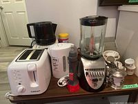 Photo showing white Cuisinart toaster, white electric kettle, GE blender, silver sugar bowl, and demitasse coffee cups with saucers on a wooden table.