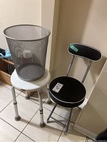 Photo shows three items grouped together: a gray wire mesh waste basket on top of a white adjustable shower stool and a black folding stool next to them.