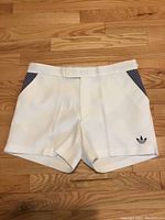 Full front view of white Adidas golf shorts with blue patterned side detail on hardwood floor.