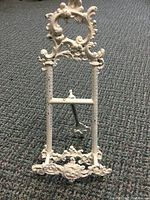 Close-up of antique white painted easel showing ornate scroll and floral decoration on upper portion and center support bar.