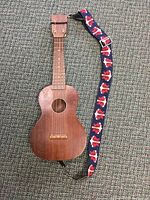 Full view of the small wooden ukulele with attached colorful strap lying on a carpeted floor.
