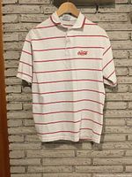 Full front view of white short-sleeve polo shirt with thin red horizontal stripes, hanging on wood hanger against brick wall background.