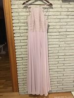 Full view of light pink dress hanging on wooden hanger against brick wall. The dress has a lace bodice and long chiffon skirt.