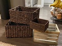 Four storage baskets including three dark brown woven wicker baskets and one wooden slatted crate style basket on a floor.