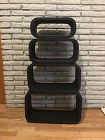 Four black wood stacking cubes arranged vertically against a white brick wall and hardwood floor.