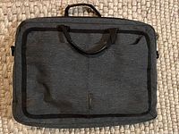 Front view of a black laptop bag lying flat on beige carpet, showing two carrying handles and a front zippered pocket.