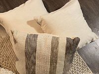 Three decorative pillows grouped together on a floor; two plain beige and one striped textured pillow in neutral shades.