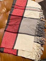 Folded plaid throw blanket on leather surface, showing large red, gray, cream, and black blocks with fringe along one side.