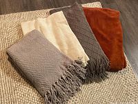 Four folded throw blankets in earth tones arranged on a woven rug, showing colors and fringe detail.