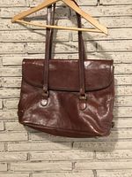 Full front view of vintage dark brown leather shoulder bag hanging on a wooden hanger against a white brick wall.