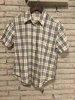 Front view of the Burberry's check patterned short sleeve button-up shirt on a wooden hanger against a brick wall.