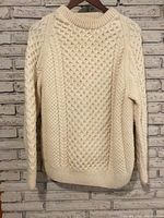 Front view of cream cable knit wool sweater hanging on a wooden hanger against a brick wall.