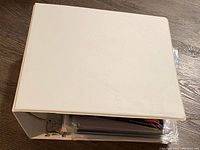 Closed white 3-ring binder/folder on a wooden floor with visible inner contents through the side opening.