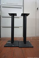Full view of two black steel stereo speaker stands showing vertical support tubes and rectangular solid bases sized approximately 2ft by 10in.