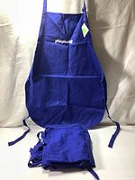 Front view of one navy blue apron showing two large pockets, adjustable neck strap, and Playmobil logo embroidered on chest area. Aprons are new, folded, and stacked beneath it.